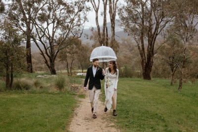 A young man wears a suit and a young bride wears a long white dress and blundstone boots, both sharing an umbrella and walking on a dirt path in a national park