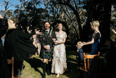 A groom wearing a kilt and a bride wearing a vintage wedding dress both exit their wedding ceremony with leaf confetti being thrown on them in celebration