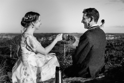 A bride wearing a vintage wedding dress and a groom wearing a kilt, both share a cup of tea sitting on rocks at the Blue Mountains, with personalised mugs