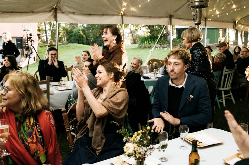 A crowd of wedding guests gather under a tent and clap for the bride and groom