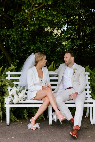 A bride and groom embrace after their garden wedding ceremony at Highwic in Newmarket - Central Auckland. The bride wears a white skirt suit and holds white flowers. The groom wears an off-white suit.