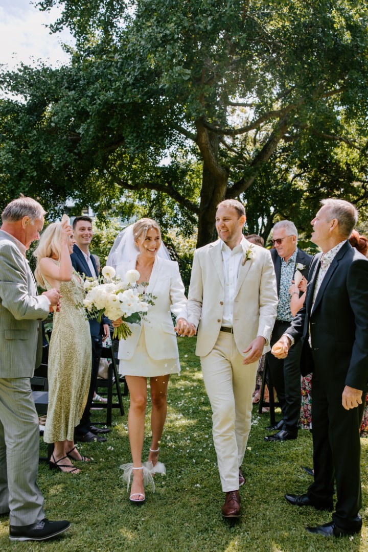 A bride and groom exit their small and intimate wedding ceremony at Highwic in Newmarket - Central Auckland. The bride wears a white suit, skirt and veil. The groom wears an off-white suit. The bride carries white flowers.
