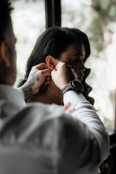 A groom helps put earrings on his bride as they get ready together in an Airbnb in Hobart Tasmania