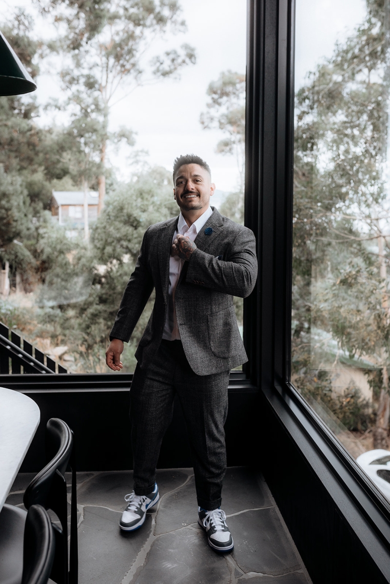A groom on his wedding day is standing adjusting his tie near a large window overlooking the beautiful bush in Hobart, Tasmania. He is wearing a suit and Nike sneakers.