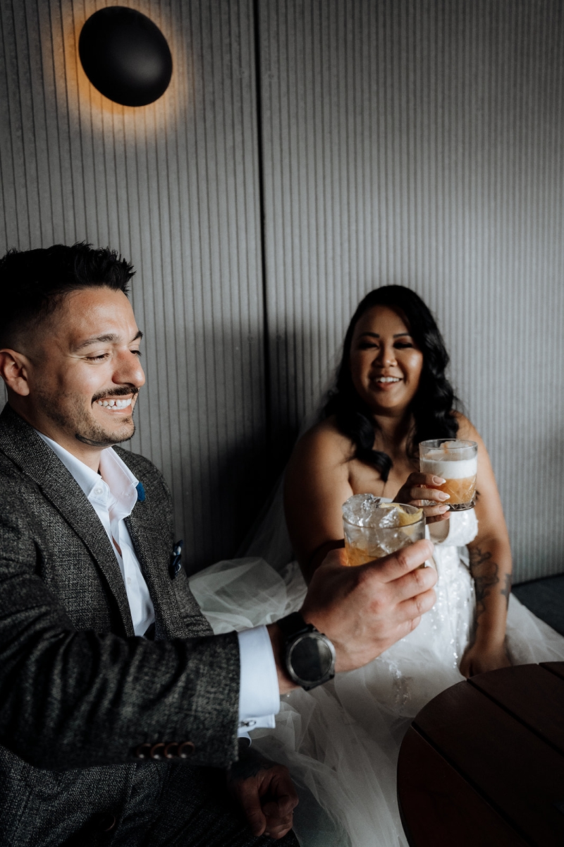 A bride and groom are dressed glamorously in wedding clothes but sit in a bar eating burgers and fries and are cheering with two drinks