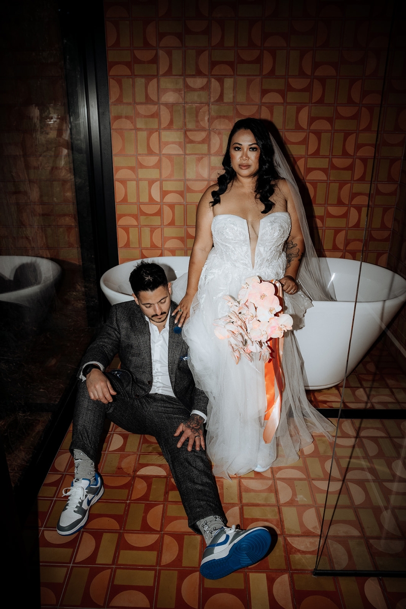 A bride and groom are dressed glamorously but sit in the retro styled bathroom of their Airbnb in Hobart, Tasmania